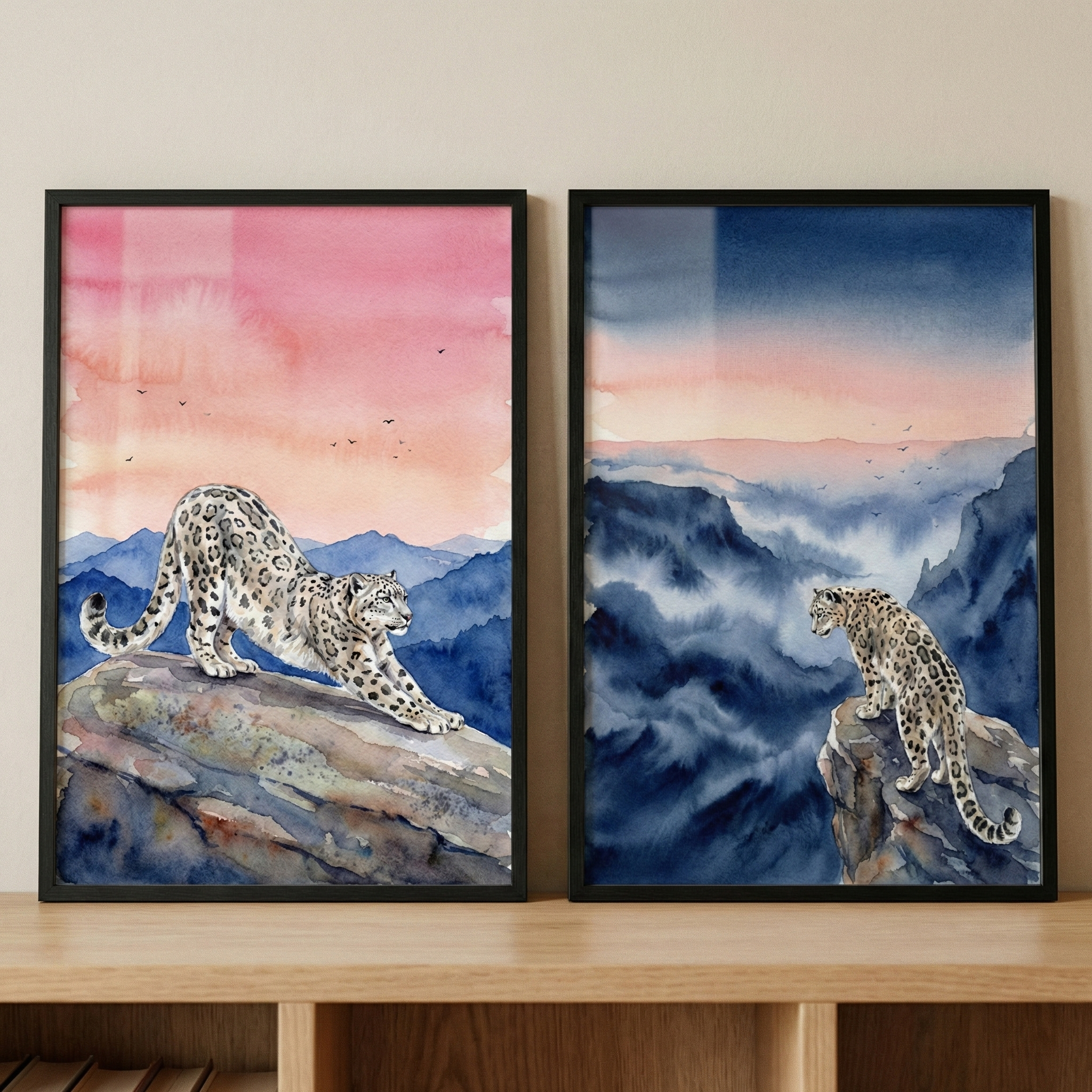 Two framed watercolor artworks sit side-by-side on a light-colored wooden shelf against a neutral wall. Both pieces depict a snow leopard in a rugged, mountainous landscape.

The artwork on the left shows a snow leopard in the foreground, stretching its body forward on a large, textured rock. Behind the animal, layers of blue mountain peaks rise under a vibrant sky that transitions from a soft orange to a deep pink, with small bird silhouettes scattered across the horizon.

