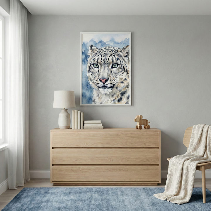 A cozy room features a light wood dresser topped with a ceramic lamp, stacked books, and a small wooden toy. Above, a striking painting of a snow leopard against a mountain backdrop hangs on the wall. A soft blue rug covers the floor, and a wooden chair draped with a cream blanket sits nearby.