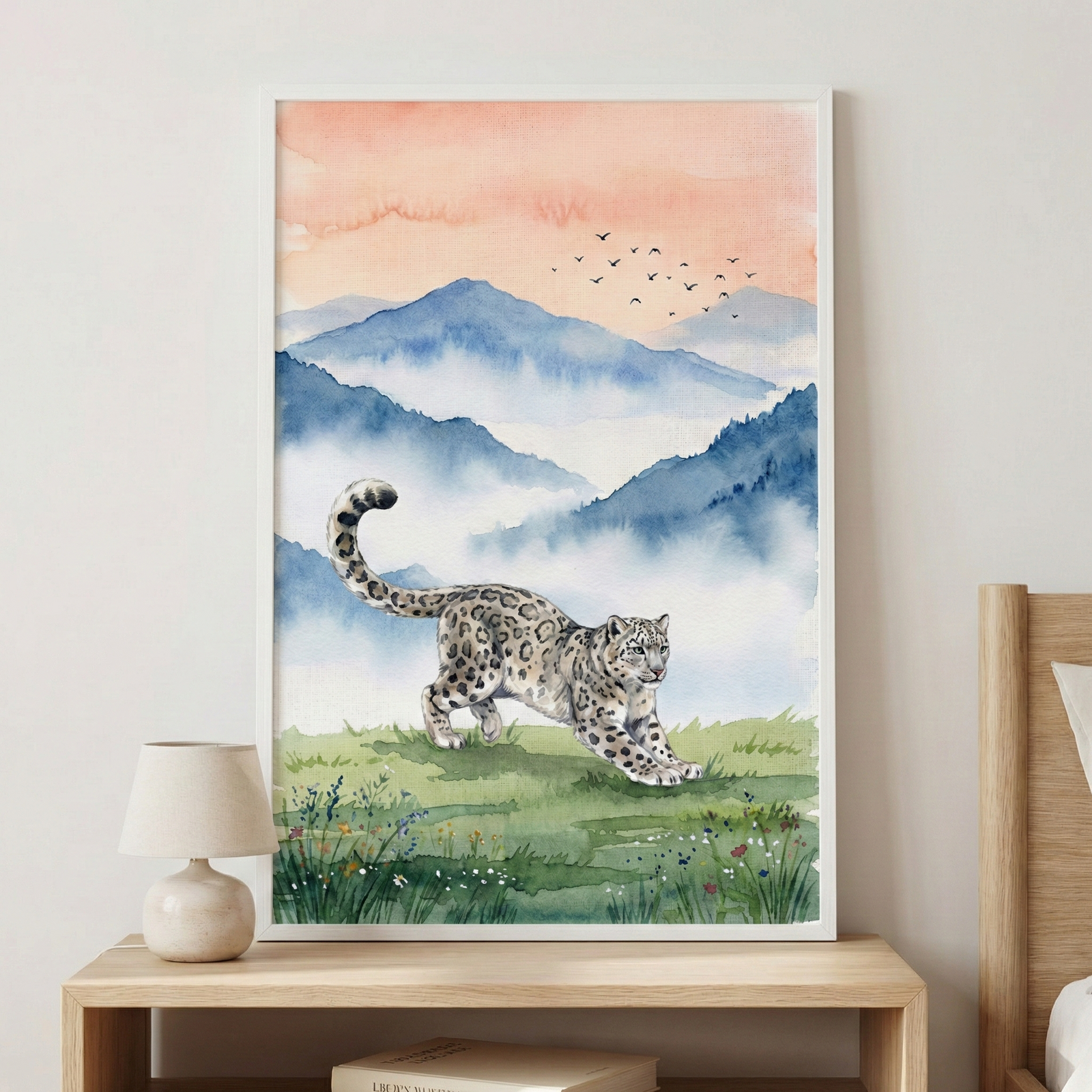 A framed piece of art sits on a light wooden table next to a small cream lamp. The illustration features a snow leopard with its distinctive spots and long tail, shown stretching across a green, flower-speckled meadow. In the background, misty blue mountains rise under a soft peach-colored sky. A small flock of birds flies in the distance. The artwork has a delicate watercolor style with soft, blended tones, creating a peaceful and serene nature scene.