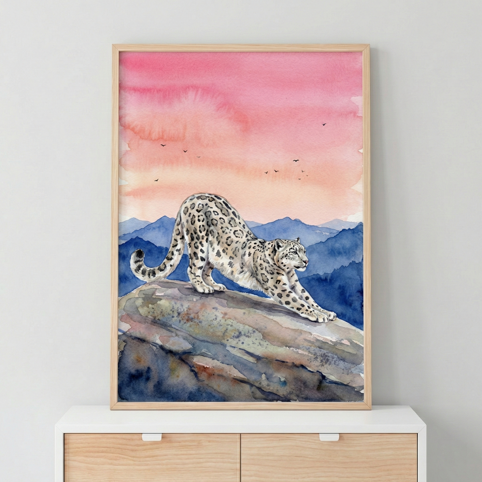 A watercolor illustration of a snow leopard stretching its body across a craggy rock. The leopard features a pale coat with dark rosettes. The background showcases layered blue mountain ranges beneath a sky of blended pink and orange hues, accented by tiny silhouetted birds. The artwork is displayed in a light-wood frame, positioned on a white surface above a wooden chest of drawers against a plain gray wall.