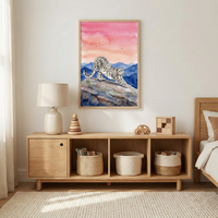 A bright, minimalist child's room features natural wood furniture against an off-white wall. A wooden sideboard holds a lamp, a wooden stacking ring toy, a toy car, and blocks. Below the surface are cubbies containing woven storage baskets. Hanging centered above is a framed illustration of a snow leopard stretching on a rocky cliff under a pink and orange sunset sky. To the right is a wooden bed frame. A cream-colored textured rug covers the floor under soft sunlight.