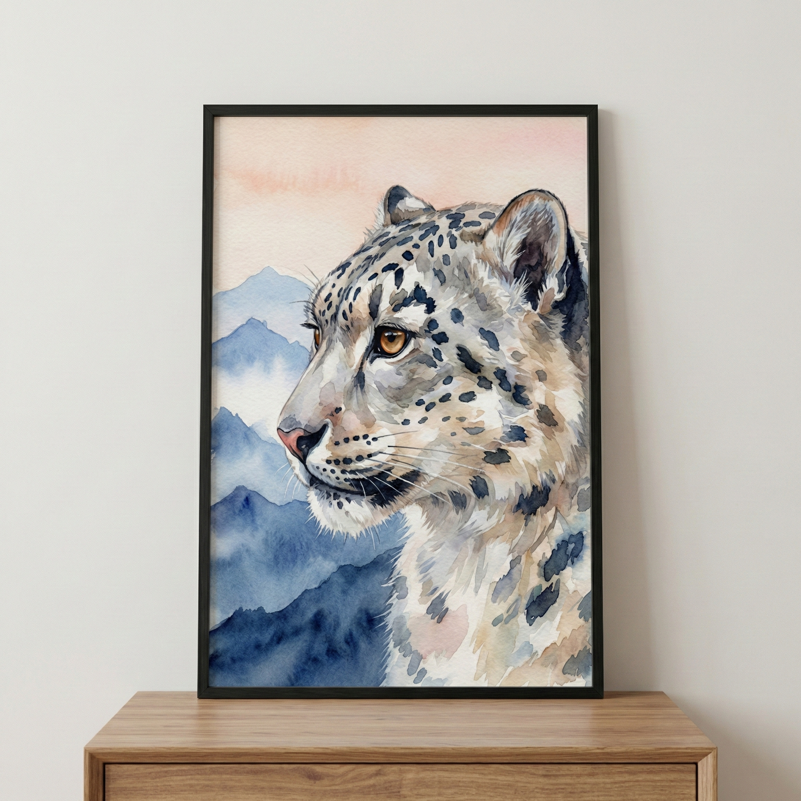 A framed watercolor-style artwork featuring the profile of a snow leopard. The animal has pale cream fur patterned with dark charcoal rosettes and a clear amber eye. The background depicts a serene mountain landscape with several layers of blue peaks fading into a soft peach-colored sky. The artwork is housed in a slim black frame, sitting on a natural wood surface in front of a neutral-toned wall.