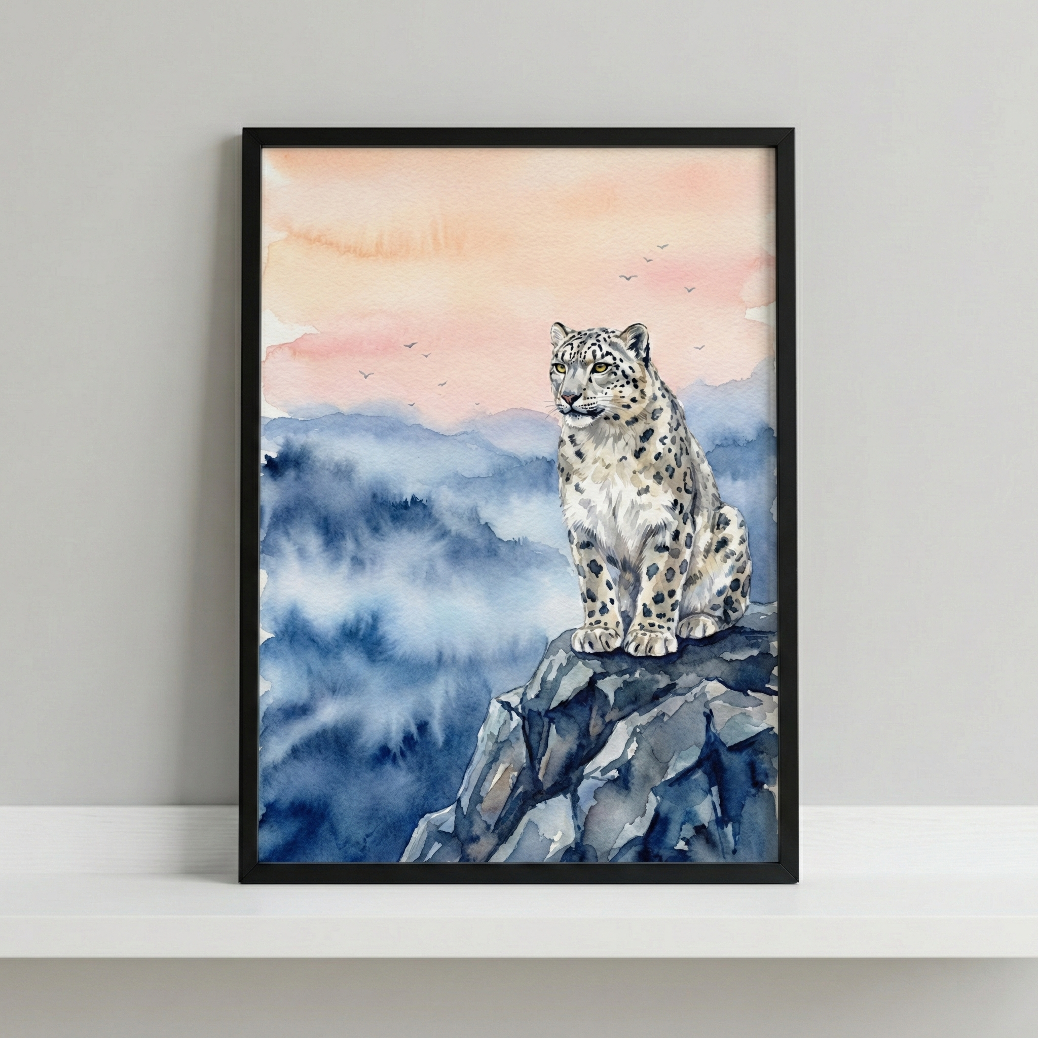 A watercolor illustration of a snow leopard sitting atop a dark, craggy rock ledge. The leopard has a pale coat with detailed dark spots and is looking toward the side. In the background, layers of misty blue mountains fade into a soft peach and light orange sky. A few small birds are silhouetted in the distance. The artwork is presented in a simple black frame, which rests on a white shelf against a neutral gray wall.