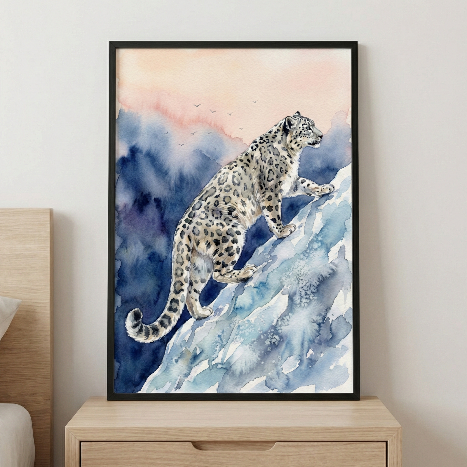 A framed watercolor illustration of a snow leopard with distinctive grey and black spots scaling a steep, icy blue mountain slope. The background consists of soft blue mountain silhouettes beneath a pale pink and orange sky where a few small birds are seen in flight. The vertical piece is held in a slim black frame and rests on a light-colored wooden nightstand. To the left, a portion of a light wood headboard and a white pillow are visible against a plain white wall.