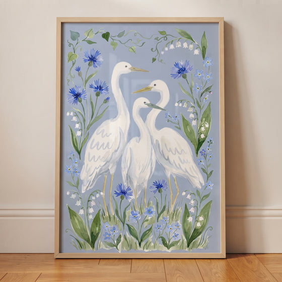 Three white egrets stand among blue cornflowers and lily of the valley in this soft blue floral egret bird wall art.