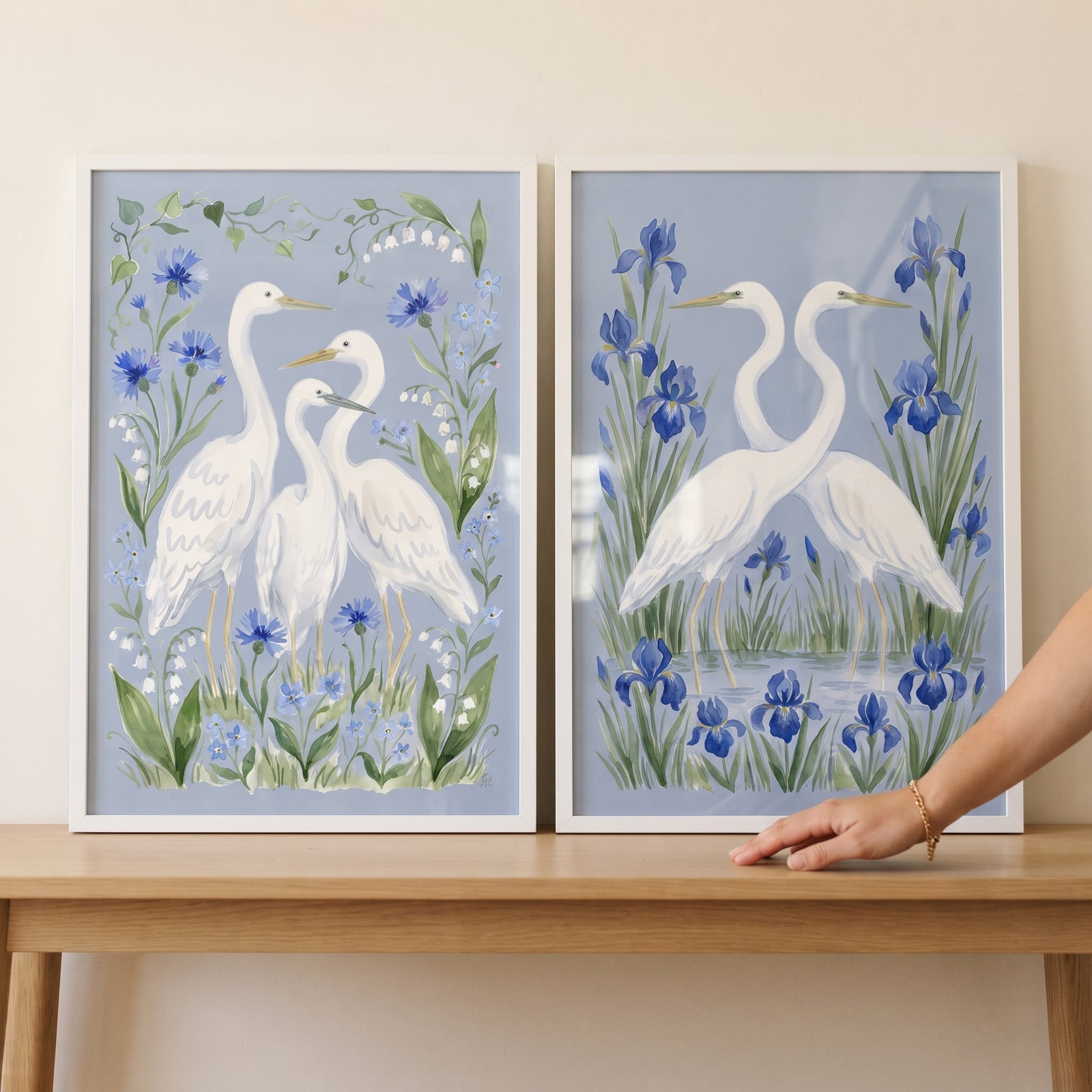 Soft blue meadow egret bird wall art set of 2 displays white egrets with blue flowers in frames on a wooden table.