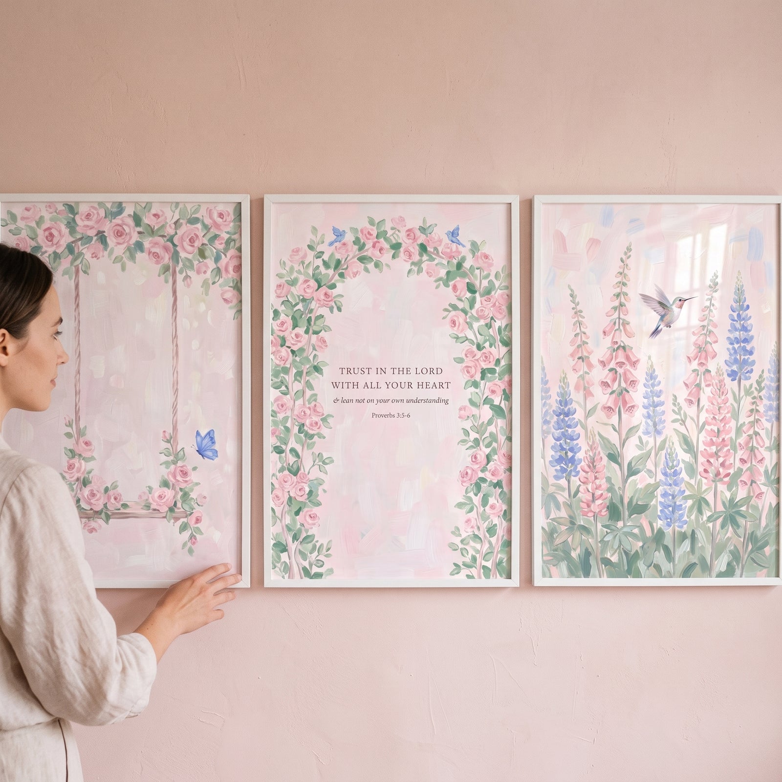 Woman hangs a soft blush botanical garden wall art set of 3 featuring floral illustrations and a Bible verse on a pink wall.