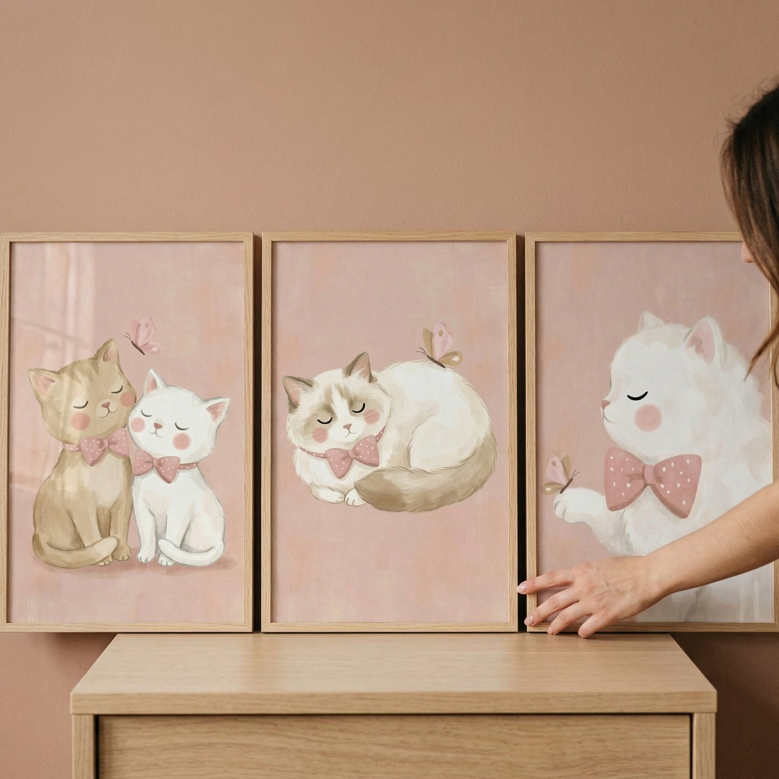 A set of three framed art prints is displayed side-by-side on a wooden surface against a solid, warm beige wall. A person's hand is visible on the right side, gently adjusting the third frame.

The artwork in each frame features cute, stylized cats in a soft, whimsical illustration style. The color palette is dominated by gentle pastel tones, particularly light pink, cream, and tan. Each illustration has a soft pink background with a subtle, textured appearance.