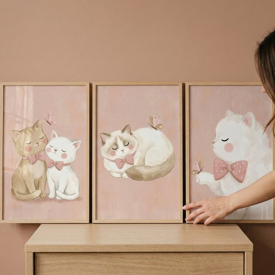 A set of three framed art prints is displayed side-by-side on a wooden surface against a solid, warm beige wall. A person's hand is visible on the right side, gently adjusting the third frame.

The artwork in each frame features cute, stylized cats in a soft, whimsical illustration style. The color palette is dominated by gentle pastel tones, particularly light pink, cream, and tan. Each illustration has a soft pink background with a subtle, textured appearance.
