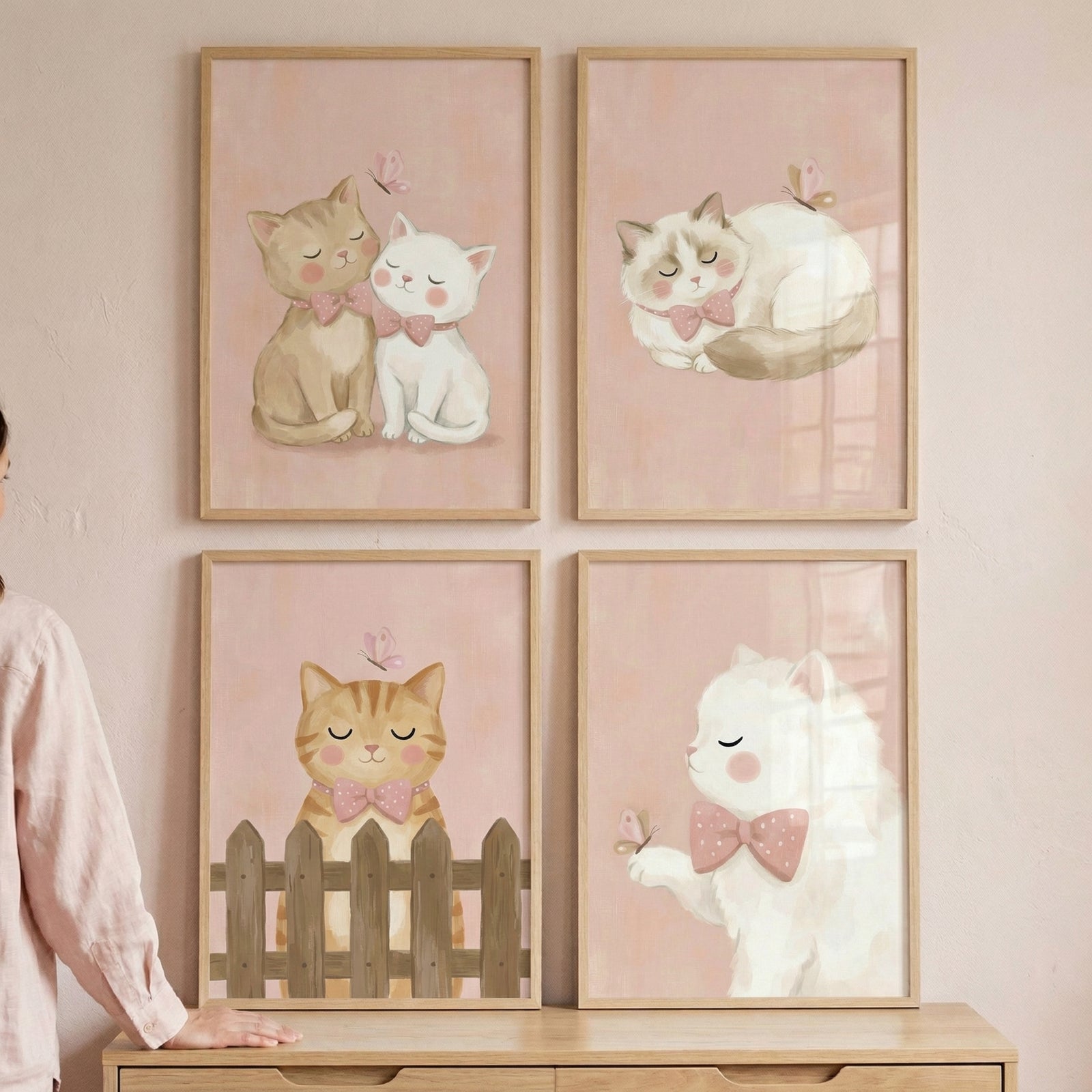 A set of four framed art pieces is displayed in a 2x2 grid on a light-colored wall. Each illustration features a whimsical, soft-colored depiction of cats with a watercolor-like texture. The background of each piece is a gentle, dusty pink hue, and all four are encased in light, natural wood frames.
