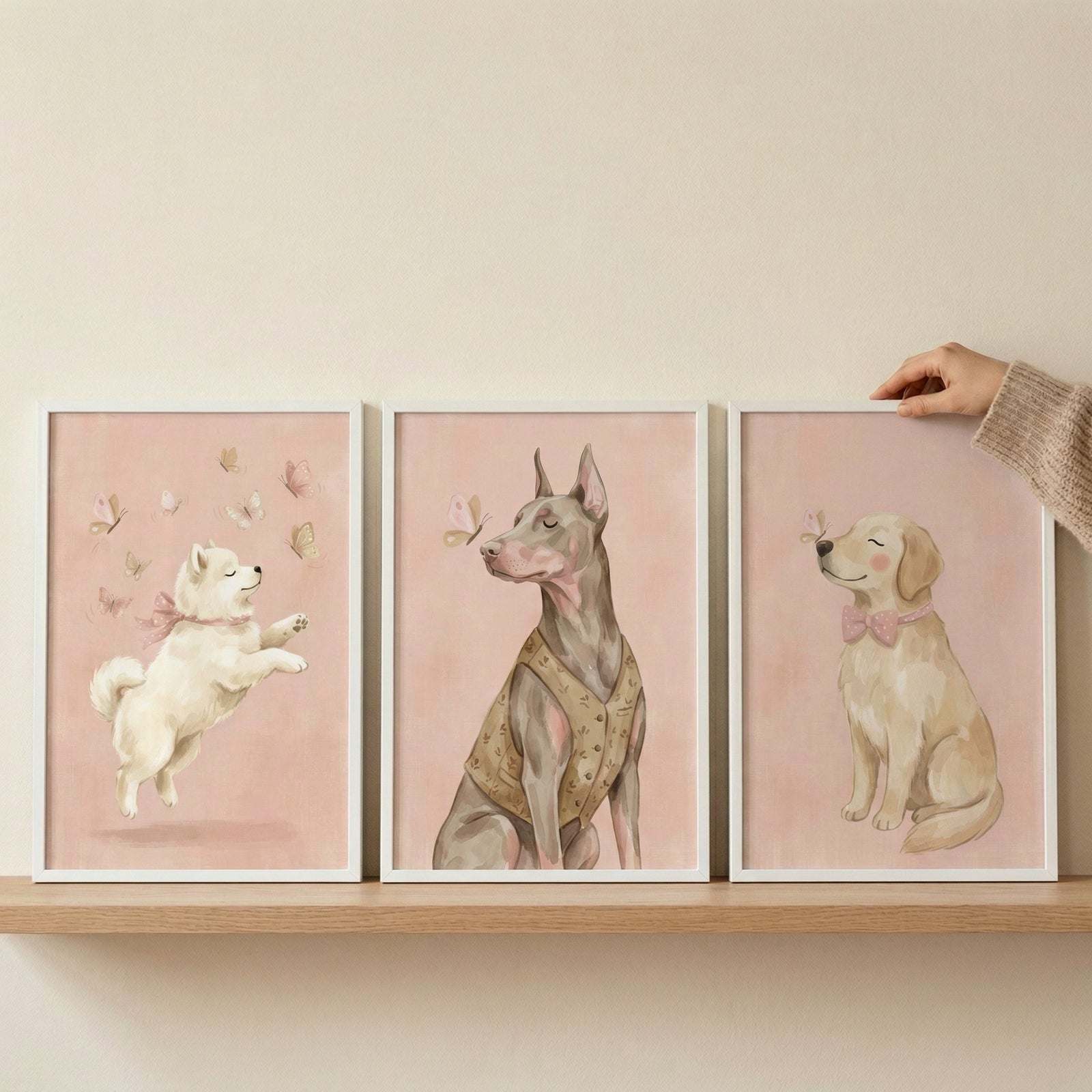 Three white-framed digital illustrations are displayed on a light wood shelf against a neutral wall. A person's hand, partially visible and wearing a beige knit sweater, is shown adjusting the frame on the right.
