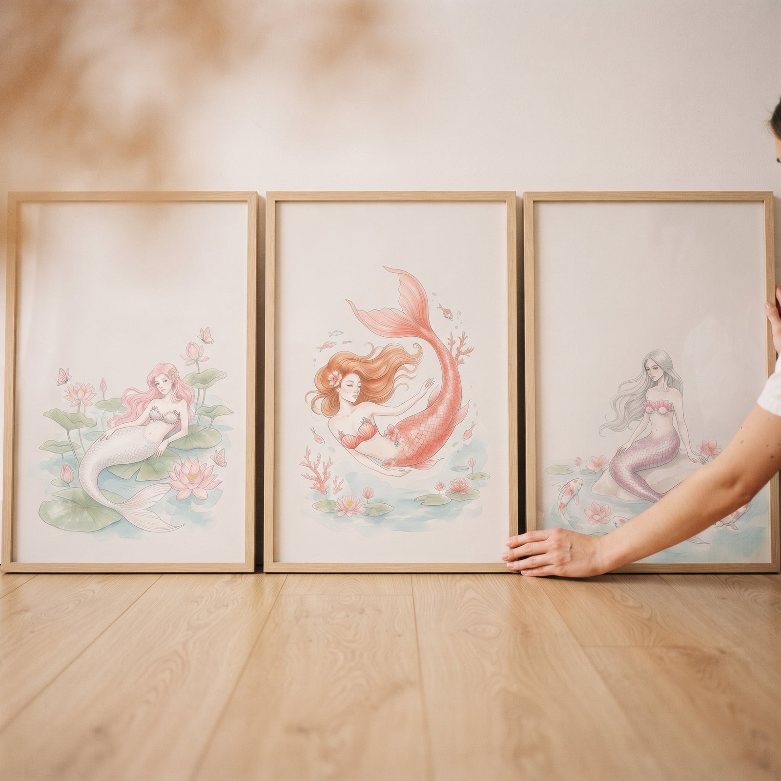 Soft blush ethereal mermaid wall art set of 3 framed prints on a wood floor as a hand adjusts the rightmost frame.