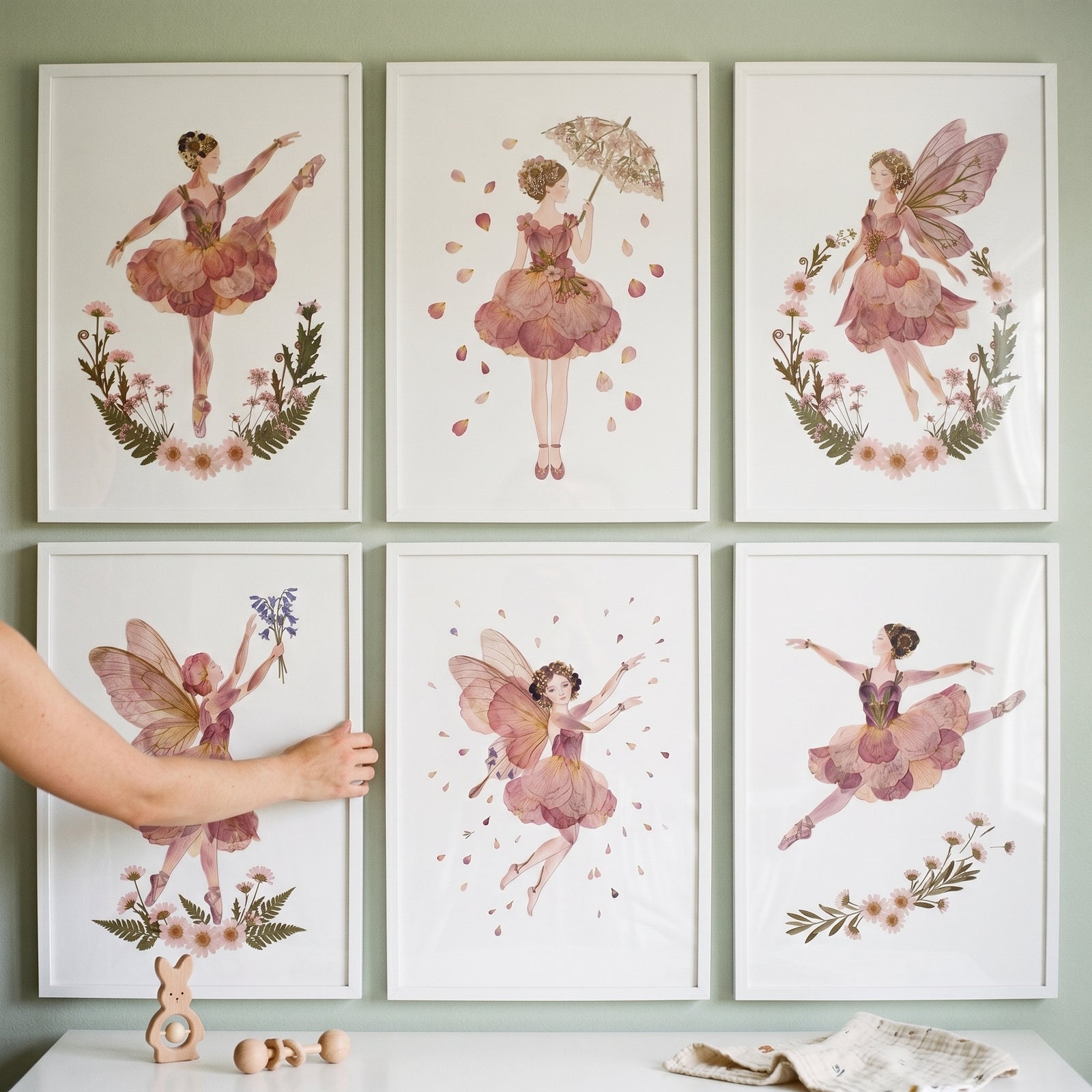 Hand adjusts a soft blush petal girl wall art set of 6 featuring fairy and ballerina illustrations in white frames.