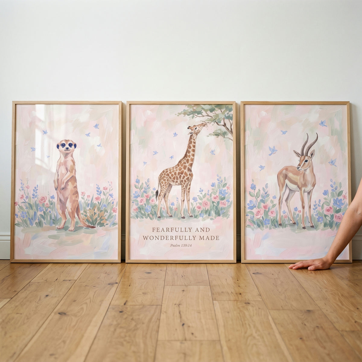 Soft blush safari animals wall art set of 3 with a meerkat, giraffe, and gazelle in floral fields leaning on a wood floor.