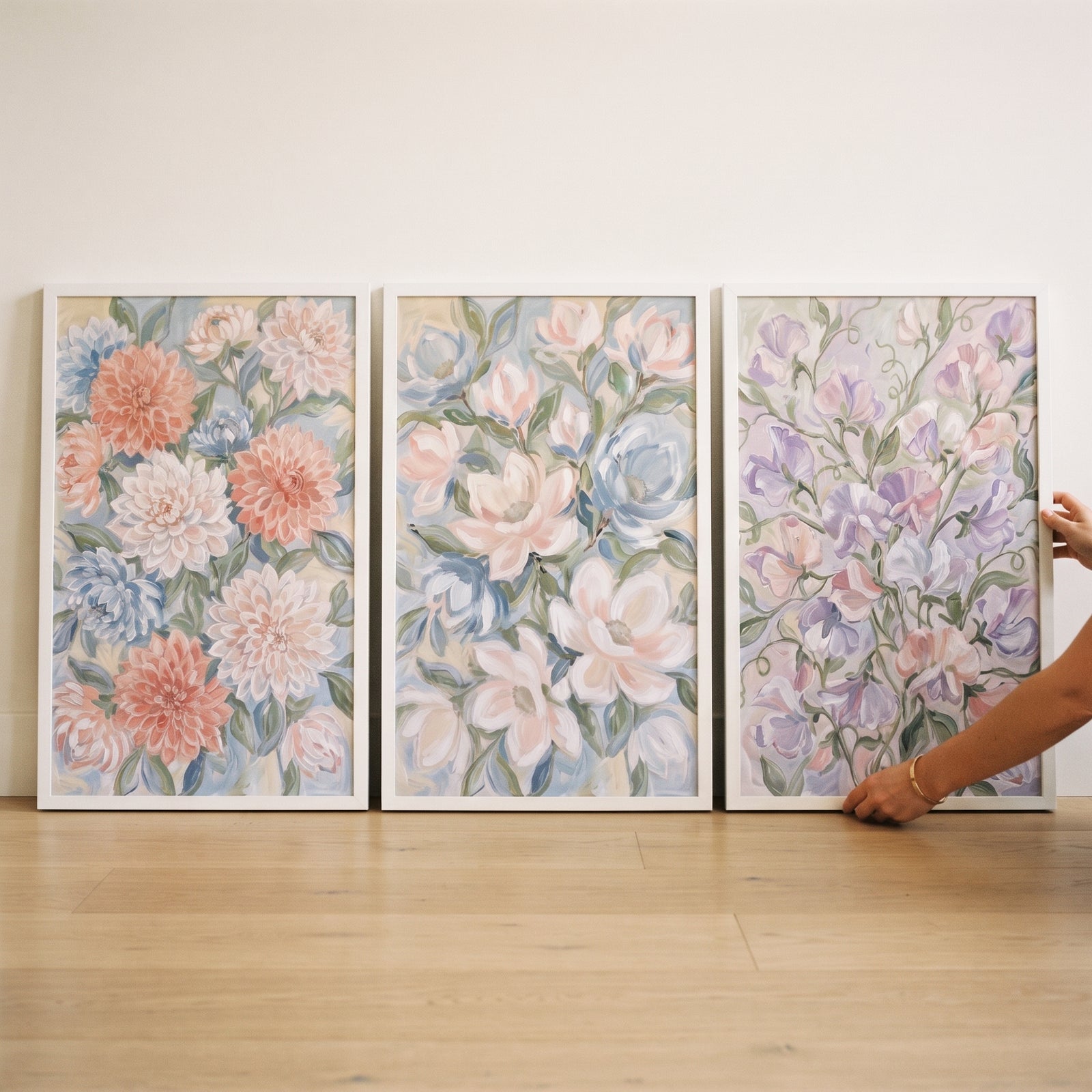 A hand adjusts a soft blush sage floral wall art set of 3 in white frames on a wood floor against a neutral wall.