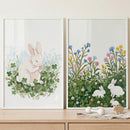 Displayed on a light wood surface against a white wall are two white-framed, textured paintings. The artwork features white bunnies in nature scenes: a single bunny sitting among green clovers on the left, and three bunnies resting in a floral meadow on the right.