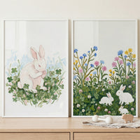 Displayed on a light wood surface against a white wall are two white-framed, textured paintings. The artwork features white bunnies in nature scenes: a single bunny sitting among green clovers on the left, and three bunnies resting in a floral meadow on the right.