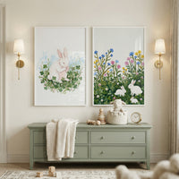 Hanging on a beige wall above a sage green dresser are two white-framed, textured paintings. The artwork features white bunnies in nature scenes: a single bunny sitting among green clovers on the left, and three bunnies resting in a floral meadow on the right.