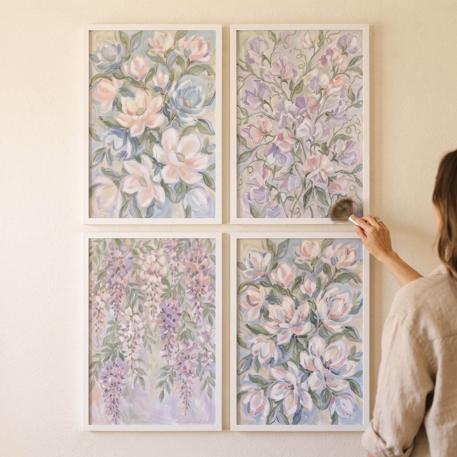 A hand holds a brush near a soft coquette garden floral artwork set of 4 hanging in white frames on a light wall.