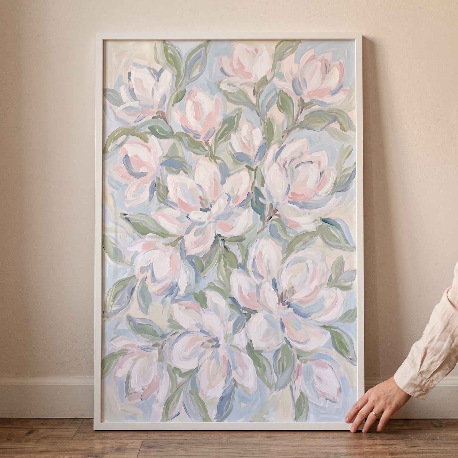 Framed soft impressionist white magnolia floral wall art leaning against a wall with a hand touching the corner.