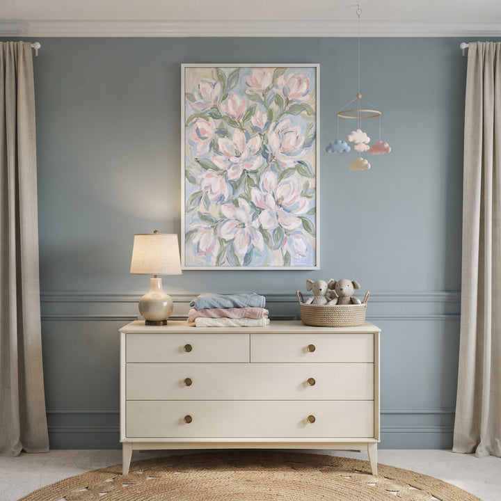 Soft impressionist white magnolia floral wall art hangs above a white dresser and lamp in a serene blue nursery.