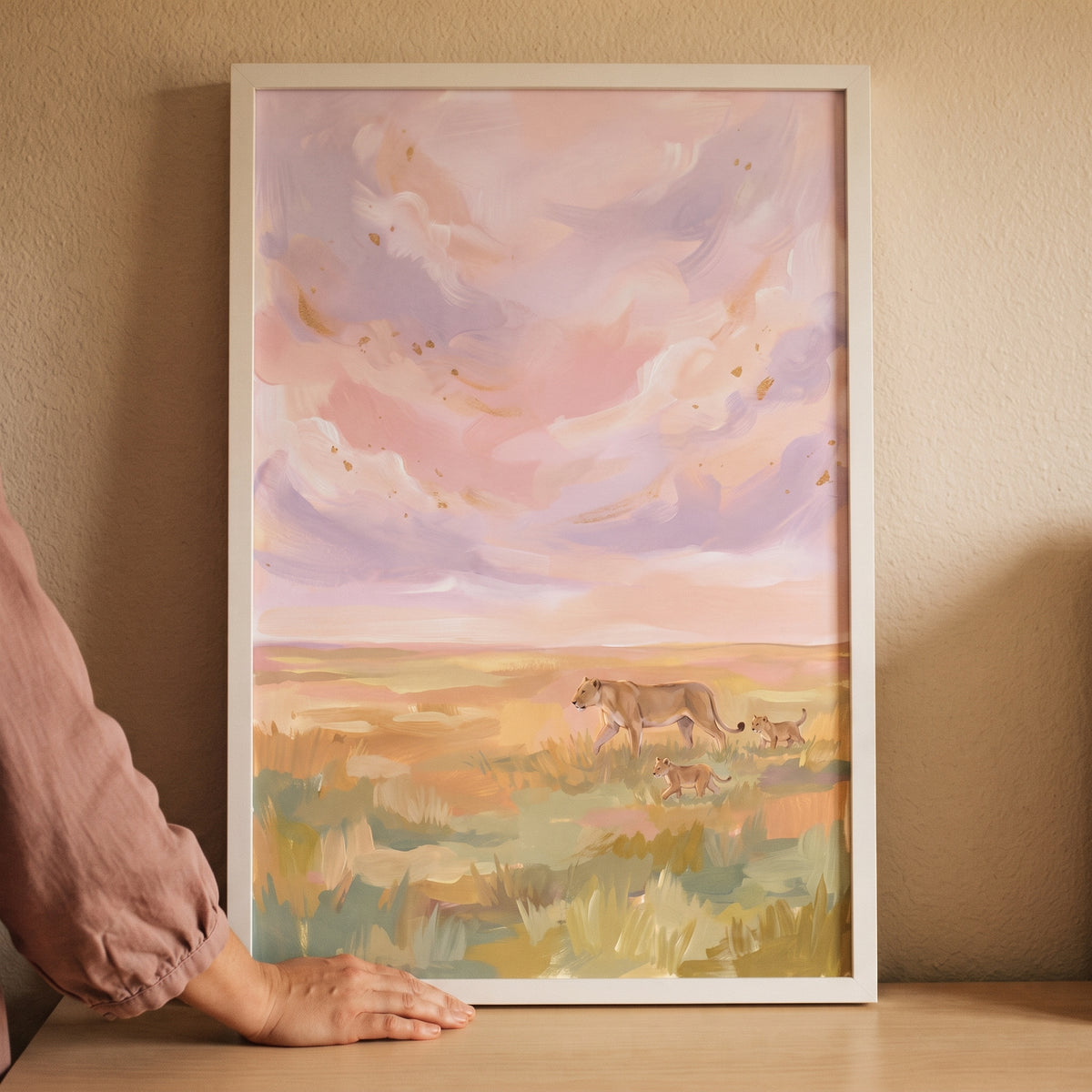 Soft lavender savanna lions wall art in a white frame touched by a hand, showing a lioness and cubs in a pastel field.