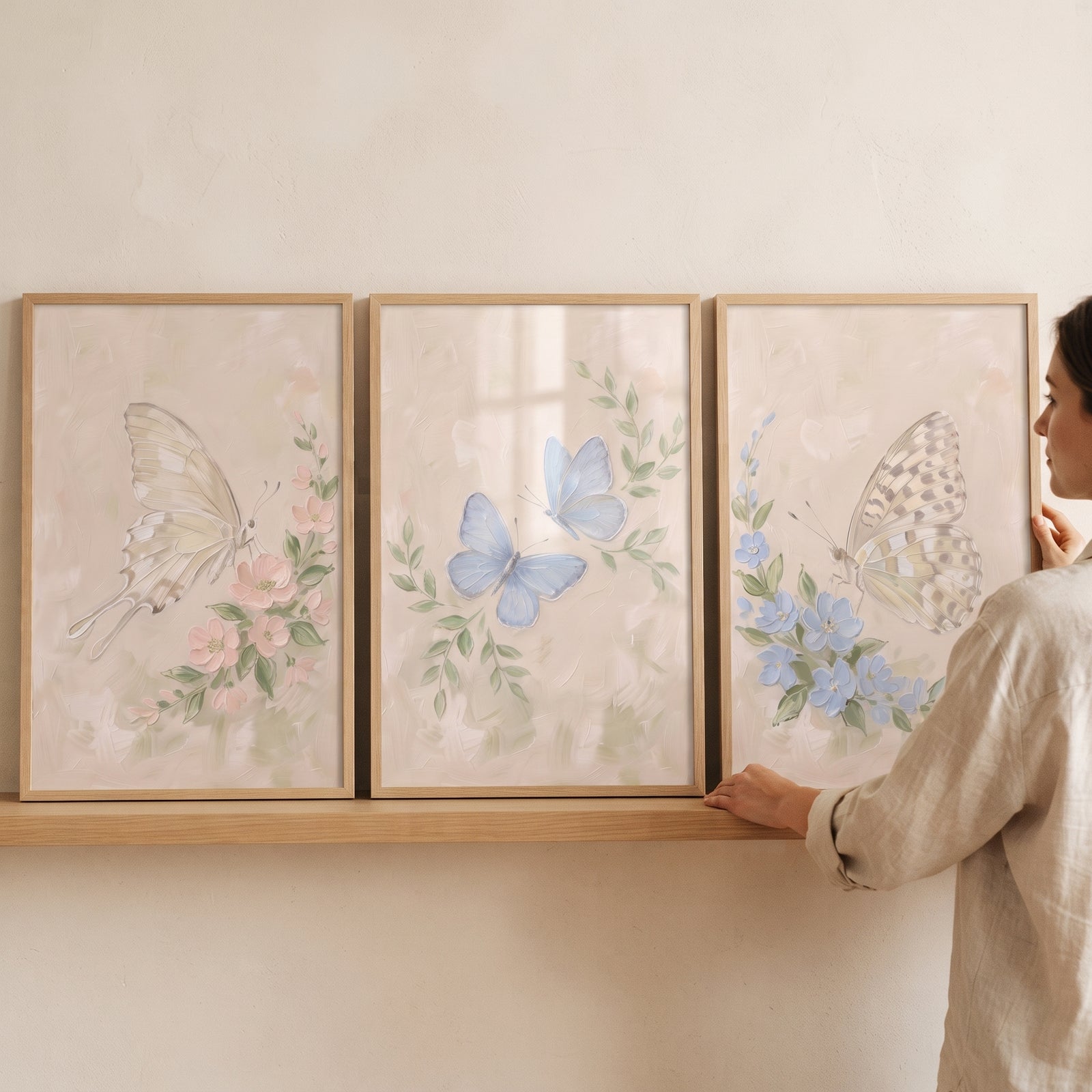Person placing soft meadow floral butterfly wall art set of 3 in light wood frames on a shelf against a beige wall.