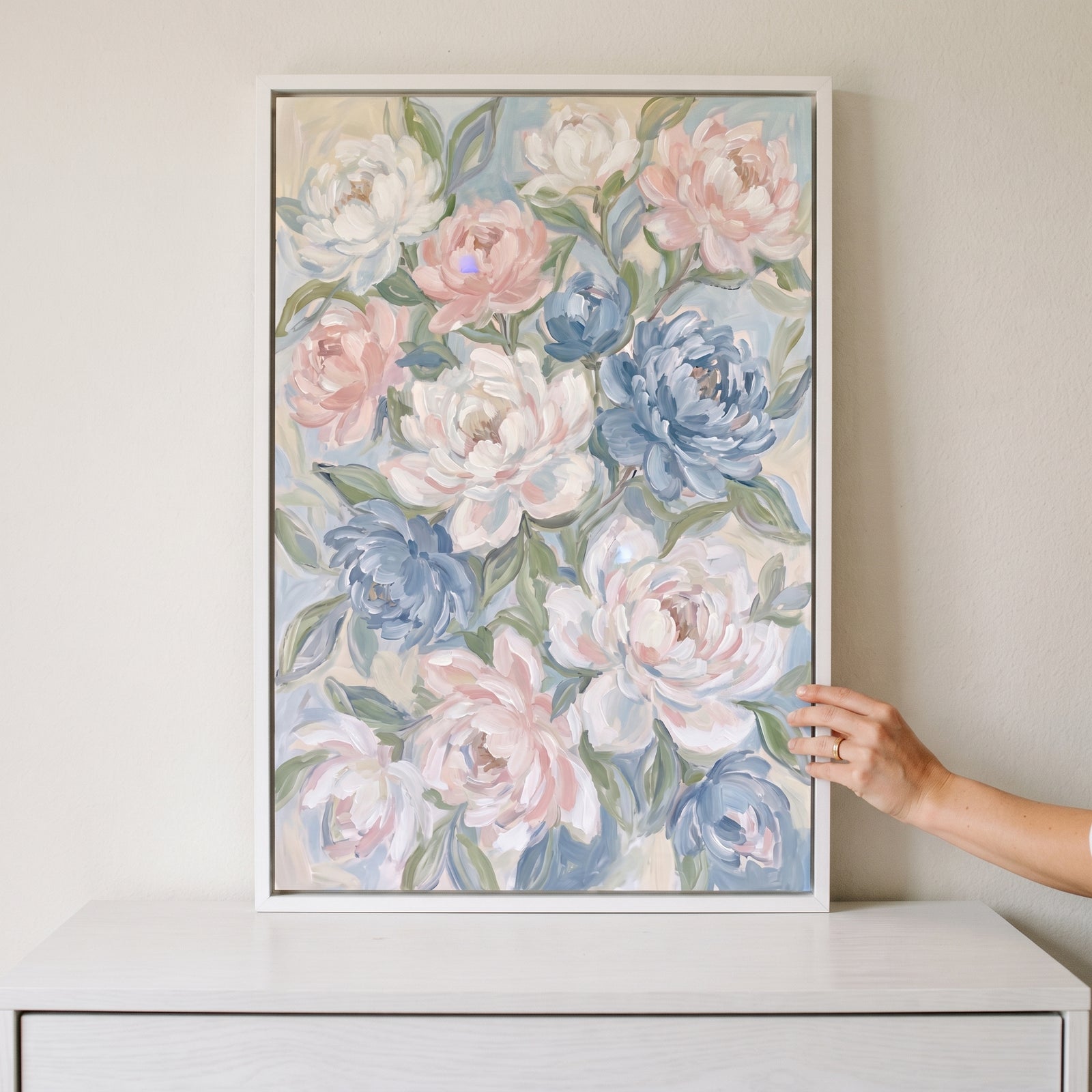 Soft painterly pink peony floral wall art in a white frame on a dresser, with a hand touching the edge.