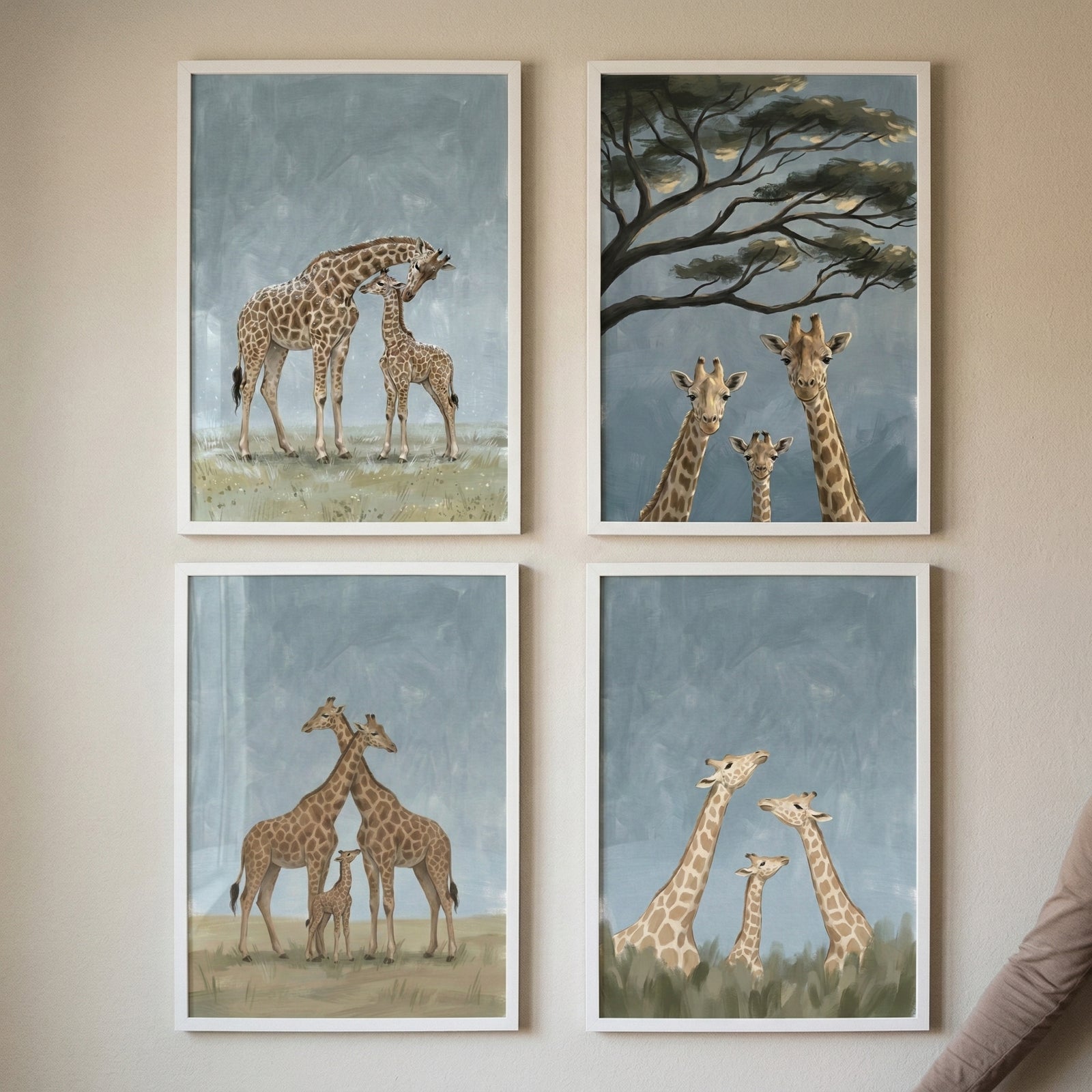 Four framed giraffe paintings arranged in a grid show gentle family moments in soft blue and neutral tones. The artworks include adult giraffes nuzzling a calf, three giraffes looking forward beneath a tree, two adults standing protectively beside a baby giraffe, and a small giraffe between two adults rising above tall grass.