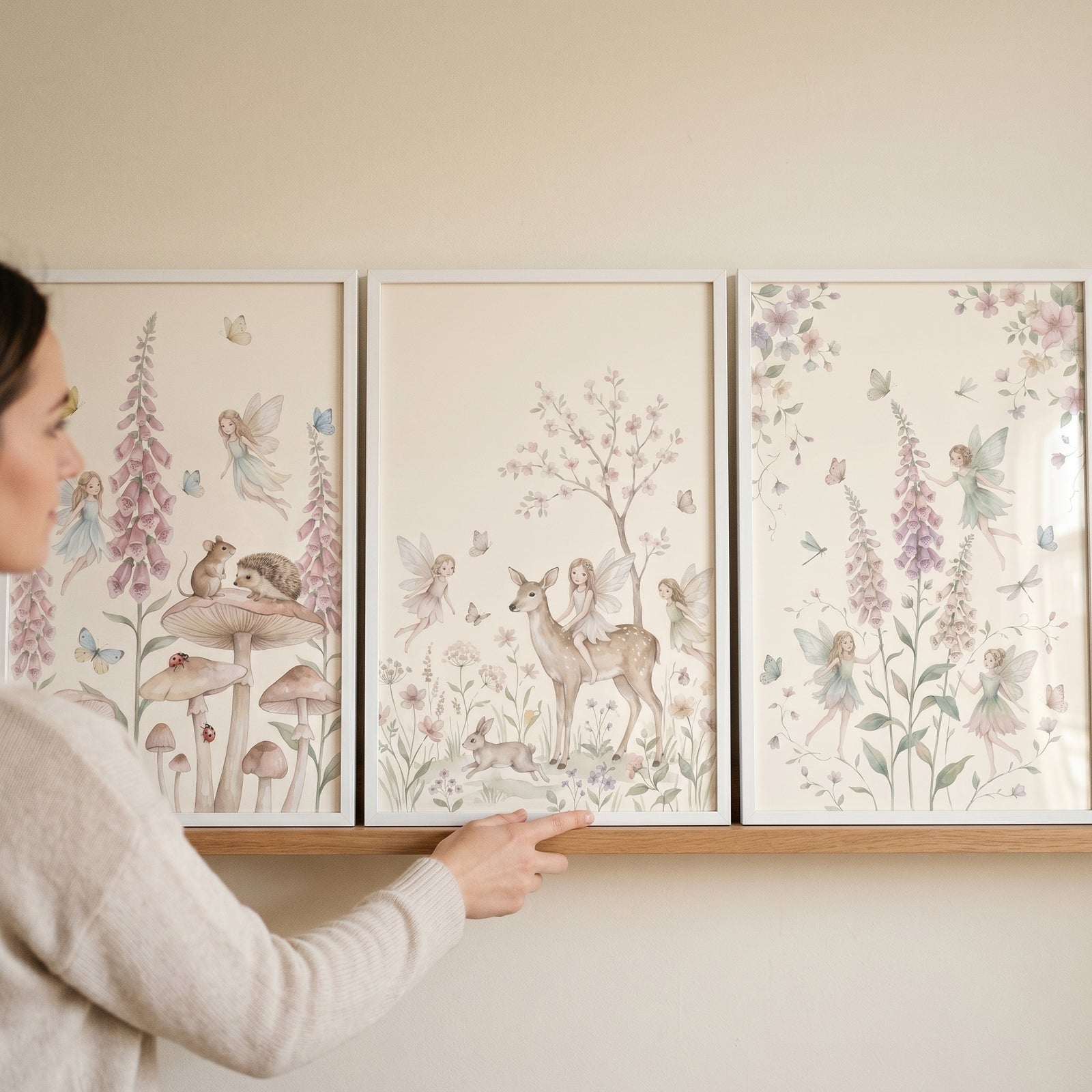 A woman's hand adjusts a soft pastel woodland fairies wall art set of 3 featuring deer and mushrooms on a shelf.