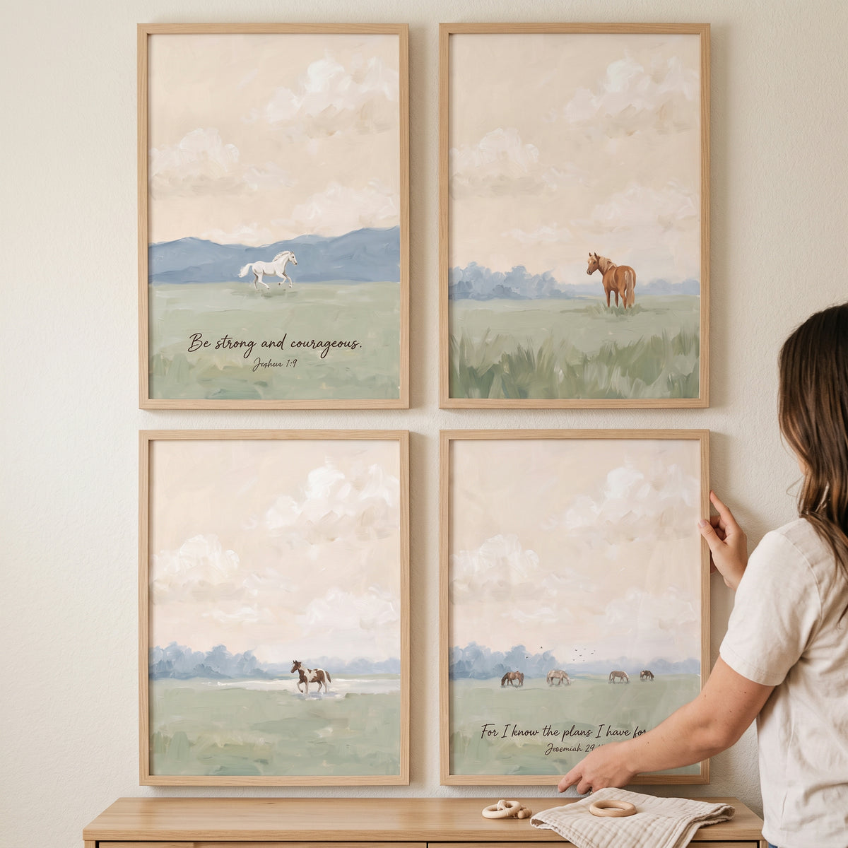 A person hangs a soft pastoral landscape horses artwork set of 4 in wooden frames on a neutral wall over a wooden cabinet.