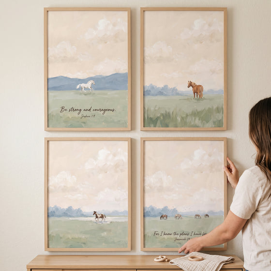 A person hangs a soft pastoral landscape horses artwork set of 4 in wooden frames on a neutral wall over a wooden cabinet.