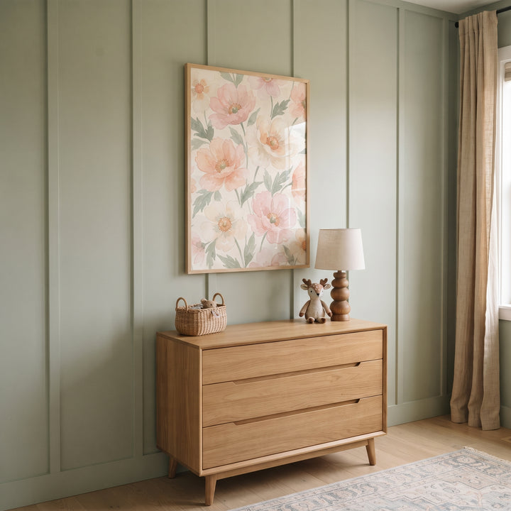 Soft peach watercolor floral wall art hangs over a wood dresser on a sage green paneled wall with a lamp and basket.