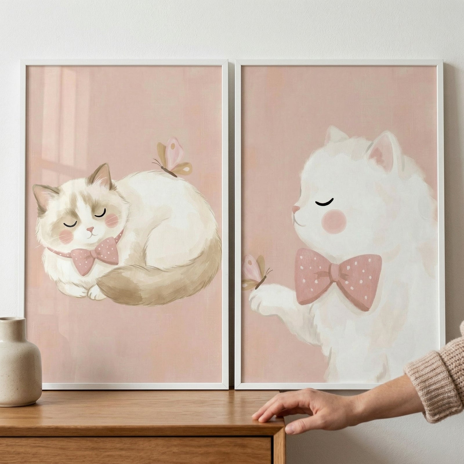 Two framed wall art pieces are positioned side-by-side on a wooden surface against a plain wall. Both illustrations feature a soft, dusty pink background. 

The piece on the left shows a fluffy, cream-colored cat with light brown markings on its head and tail. The cat is curled up as if sleeping, with its eyes closed and a small pink polka-dot bow tie around its neck. A tiny, light-pink butterfly is perched on its back. 
