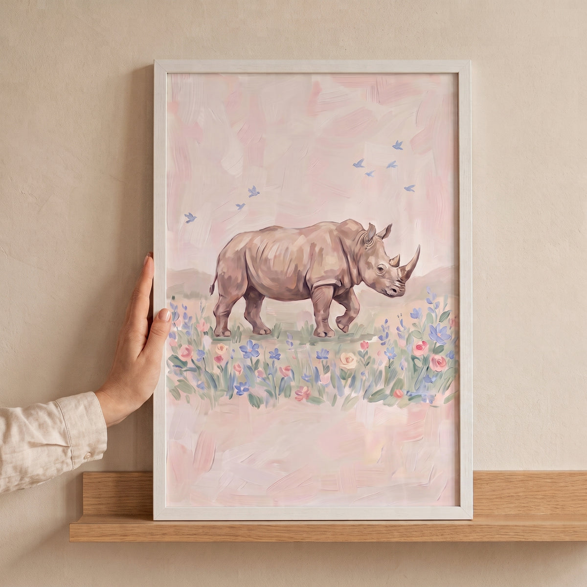 Hand holds white framed soft pink rhino floral wall art depicting a rhino and birds in a meadow on a wooden shelf.