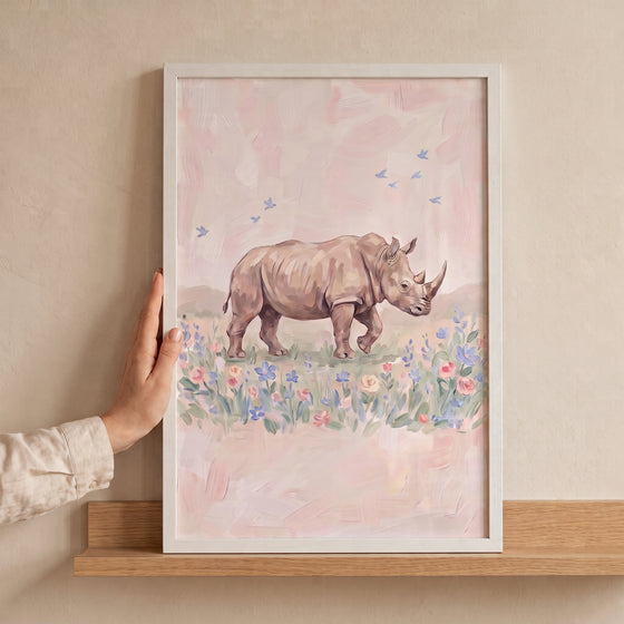 Hand holds white framed soft pink rhino floral wall art depicting a rhino and birds in a meadow on a wooden shelf.