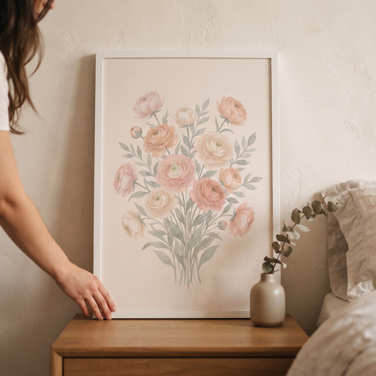 A hand touches a framed soft pink watercolor floral wall art piece on a wooden nightstand next to a bed.