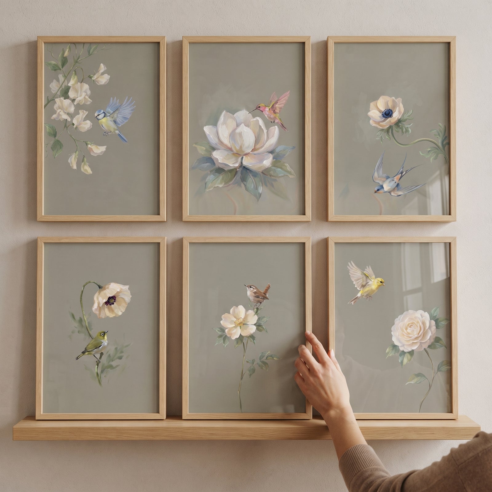 Soft sage botanical bird wall art set of 6 in wood frames on a shelf as a hand adjusts one piece.