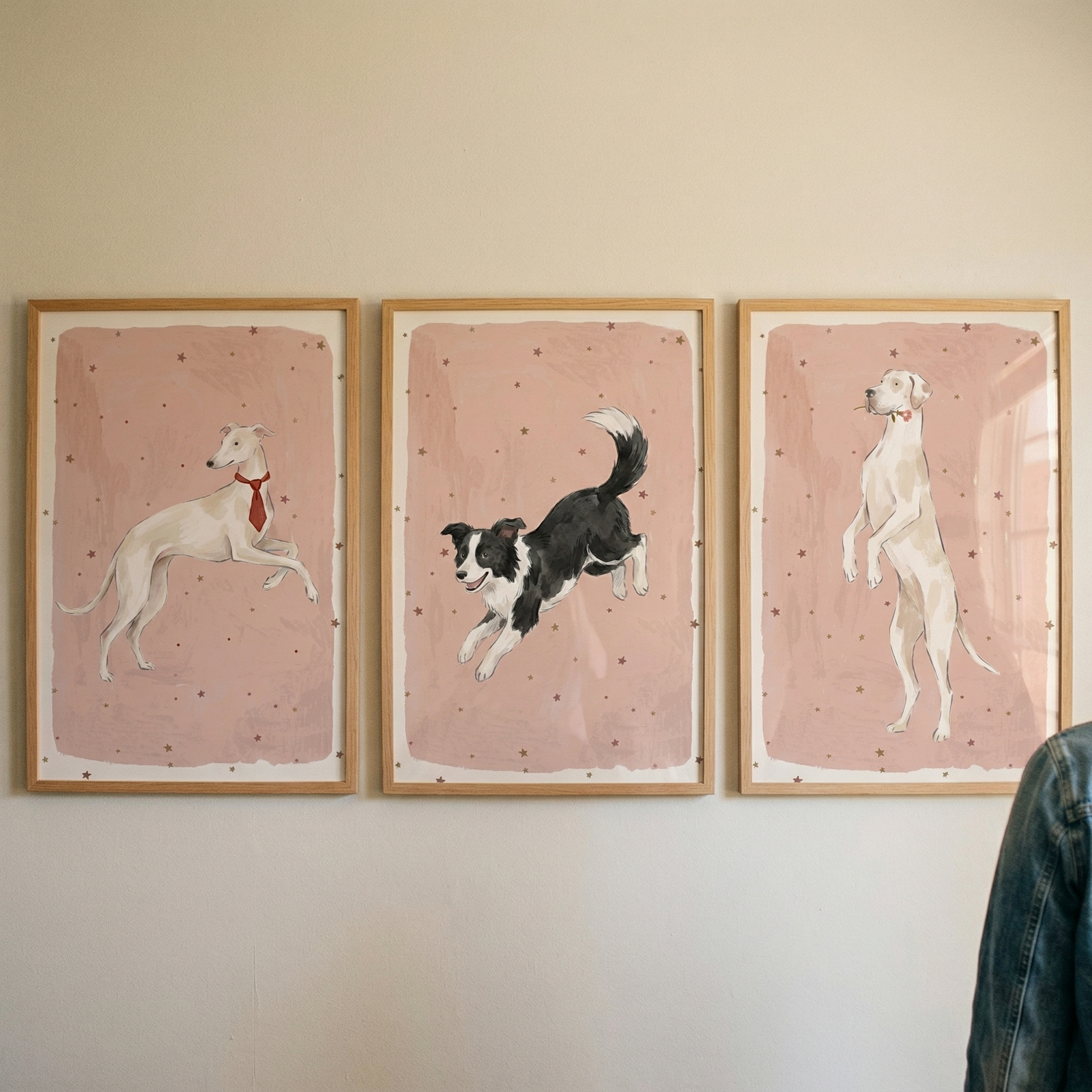 Three framed vertical illustrations of dogs hang side-by-side on a plain wall. Each piece has a dusty pink background dotted with small stars. The left frame shows a slender white dog wearing a red necktie in mid-stride. The middle frame features a black and white dog jumping through the air. 