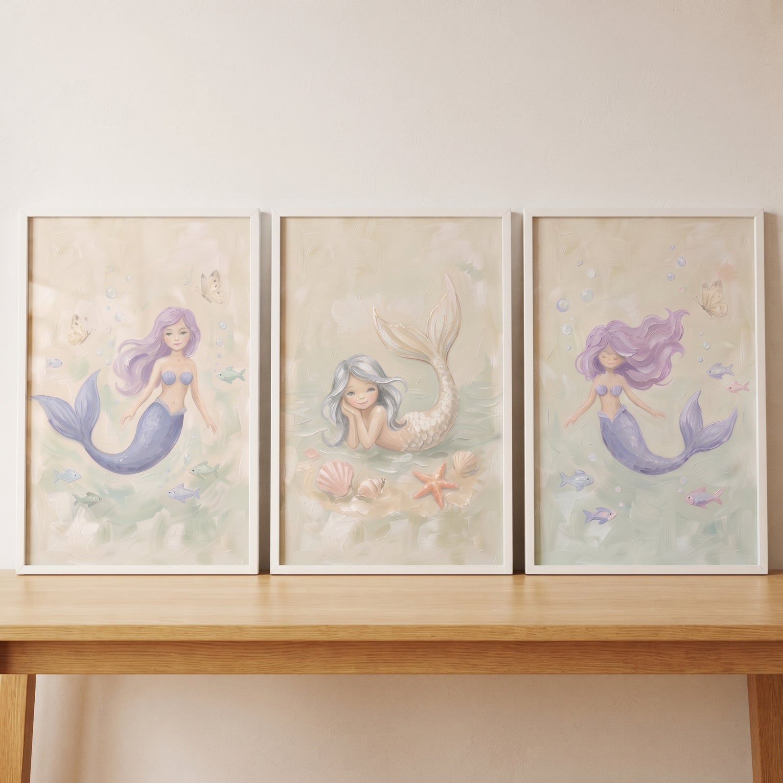 Soft subaqueous fantasy mermaid wall art set of 3 features pastel mermaids and sea life in white frames on a wood table.
