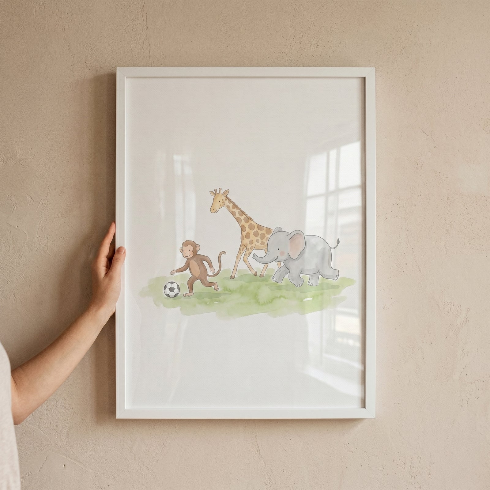 A hand holds framed soft watercolor soccer animals wall art of a monkey, giraffe, and elephant with a soccer ball.