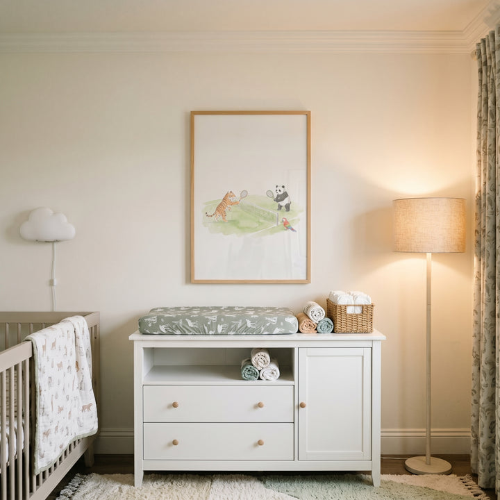 White changing table centered under soft watercolor tennis animals wall art in a nursery with a crib and floor lamp.