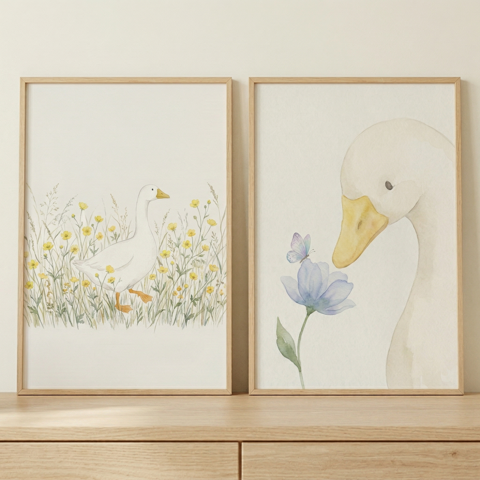 Two framed vertical watercolor-style illustrations sit side-by-side on a light wood surface against a plain wall. The frame on the left depicts a white goose walking through a field of tall grass and yellow wildflowers. The frame on the right shows a close-up of a white goose’s head as it looks down at a pale blue flower with a small butterfly resting on it. Both artworks feature a soft, muted color palette. The frames are thin and made of light-toned wood, matching the furniture below.