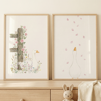 Two framed watercolor-style illustrations of white geese are displayed side-by-side on a wooden dresser. The left illustration features a goose sitting among colorful wildflowers next to an old wooden fence post with climbing pink roses. The right illustration shows a goose from behind, looking up as delicate pink petals fall around it. Below the frames, a small knitted bunny toy and a soft cream blanket sit on the light wood surface, creating a gentle and calm nursery scene.