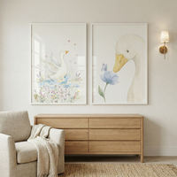 A serene interior scene showcasing two large, vertically-oriented framed illustrations on a neutral wall. The left image features a graceful white swan in water surrounded by soft pink and purple wildflowers. The right image shows a close-up of a white bird's head next to a delicate blue flower. Beneath the frames is a light-colored wood dresser. To the left, a cozy beige armchair is draped with a textured knit throw. A brass wall sconce is on the right, and a woven rug covers the floor.