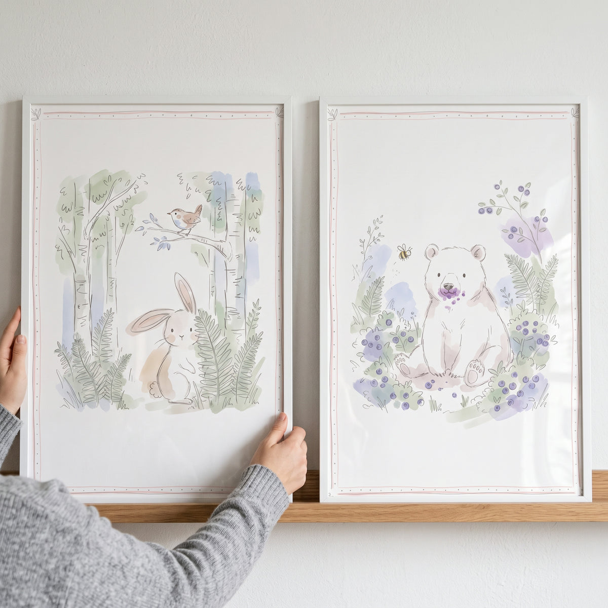 A person holds a soft watercolor woodland creature wall art set of 2 featuring a bunny, a bird, and a bear in white frames.
