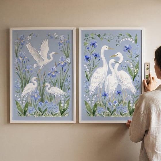 A person levels a sophisticated coastal white heron bird wall art set of 2 featuring blue flowers.