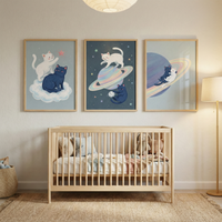 A light-filled nursery features three framed illustrations of cute cats in a space theme hanging above a wooden crib. The leftmost artwork shows a white and a dark blue cat on a cloud. The center piece depicts them near a ringed planet against a starry background. The right artwork shows the cats cuddling on planetary rings. The room has neutral decor, including a jute rug, a woven basket, a floor lamp, and a paper lantern ceiling light.