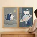 Two framed vertical artworks hang side-by-side on a plain wall. The left illustration shows a dark blue cat and a white cat leaping across a sparkling, colorful trail of light in a star-filled sky. The right illustration depicts the same two cats sitting back-to-back on a floating asteroid, their tails crossing. The background features a dark space scene with distant stars and planets. Both pieces have simple white frames. A hand in a brown sweater sleeve holds the edge of the right frame.