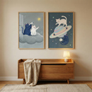 Two framed illustrations of stylized cats in space hang on a neutral-toned wall above a light-wood dresser with six drawers. Each drawer has a simple, round wooden knob. The frame on the left depicts a dark blue cat and a white cat leaping over a vibrant, multicolored cosmic trail against a dark, starry background. The frame on the right shows the same two cats sitting side-by-side on a floating rock, looking out at planets and stars. 